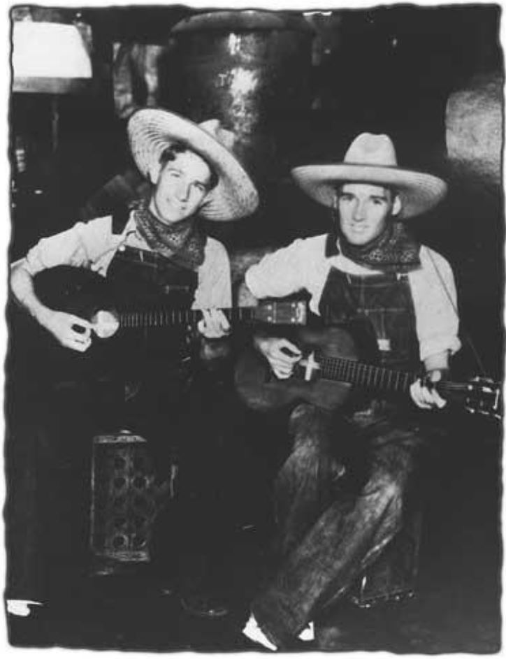 Two musicians in traditional attire sit together, playing acoustic guitars. Their outfits include wide-brimmed hats and neckerchiefs, capturing a vintage musical atmosphere.