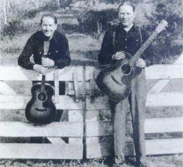 Two men stand by a wooden gate, each holding a guitar. They wear black shirts and look towards the camera, set against a rural landscape.