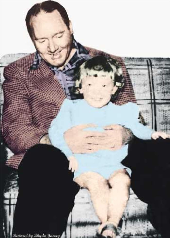 Man in a plaid jacket smiles while holding a young child in a blue outfit on a vintage couch, conveying warmth and nostalgia.