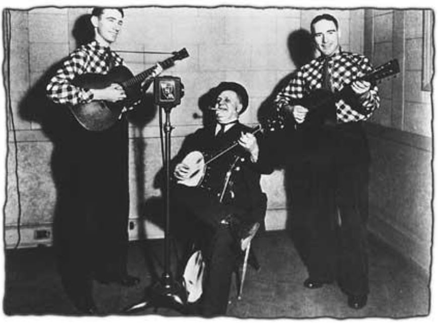 Three musicians perform together: two men play guitars while one, seated, strums a banjo. They wear checkered shirts, creating a lively atmosphere.