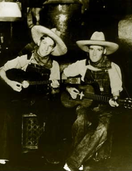 Two musicians in cowboy hats and overalls play guitars in a rustic setting, capturing a nostalgic, Western-themed musical moment.
