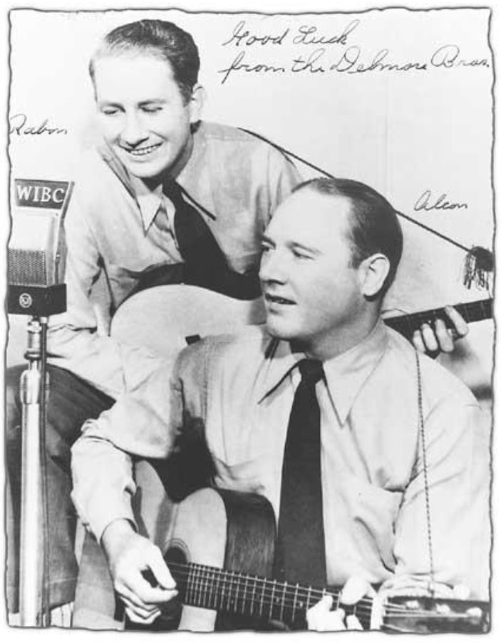 Two musicians perform on air at a radio station, one playing guitar while the other appears excited. A microphone and handwritten note are visible.