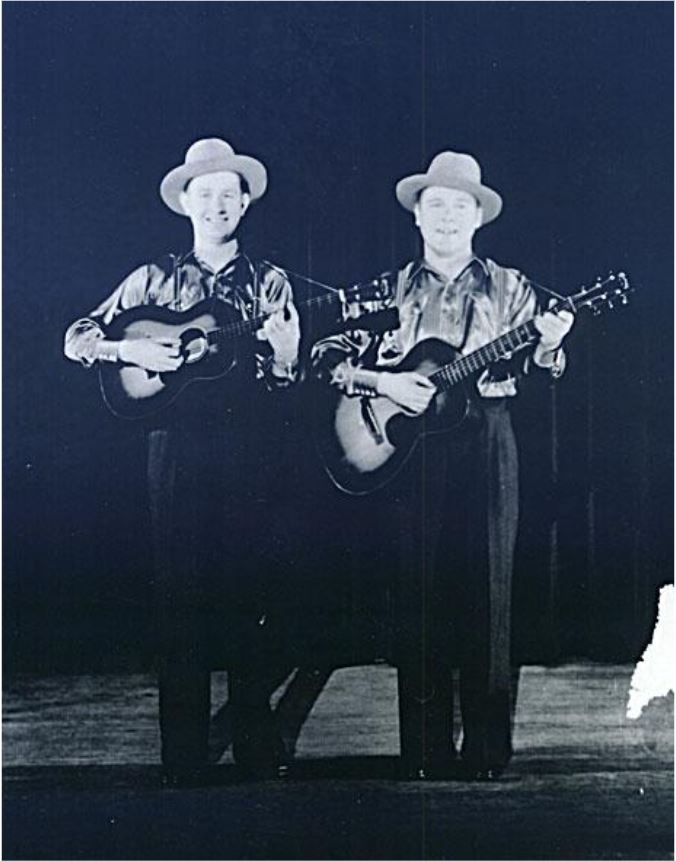 Twin musicians in matching outfits perform with guitars on stage, showcasing a nostalgic musical moment from the past.