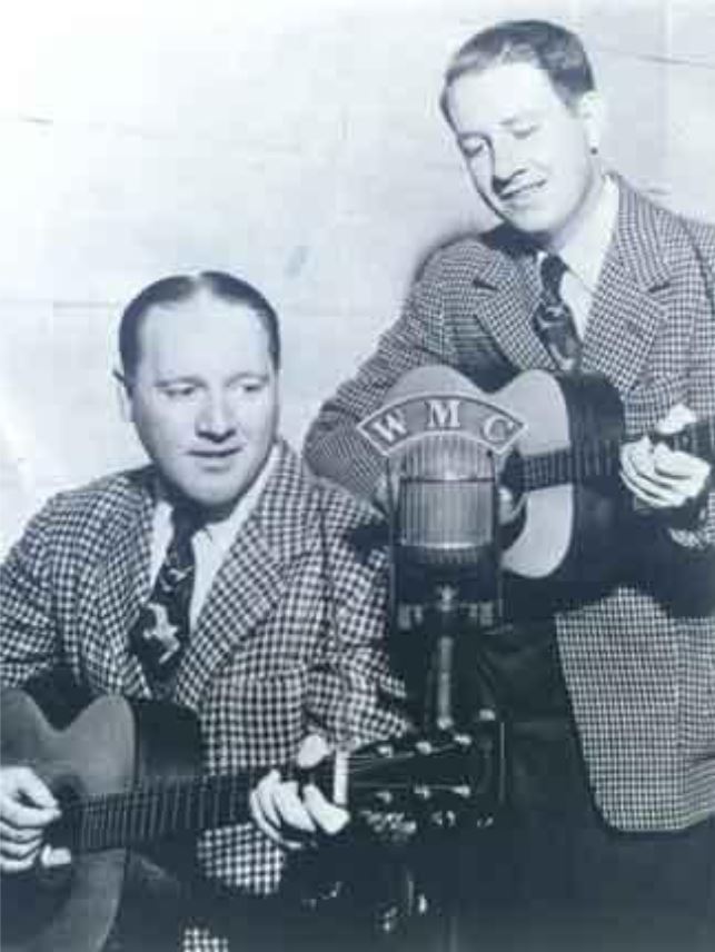Two musicians performing live with guitars near a vintage microphone labeled "WMC," showcasing a classic Americana music scene.