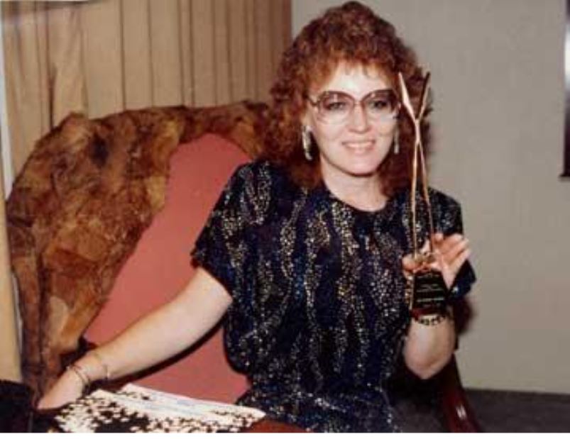 A smiling woman with curly red hair, wearing glasses and a sparkly dress, holds an award while seated at a table, showcasing her achievement.